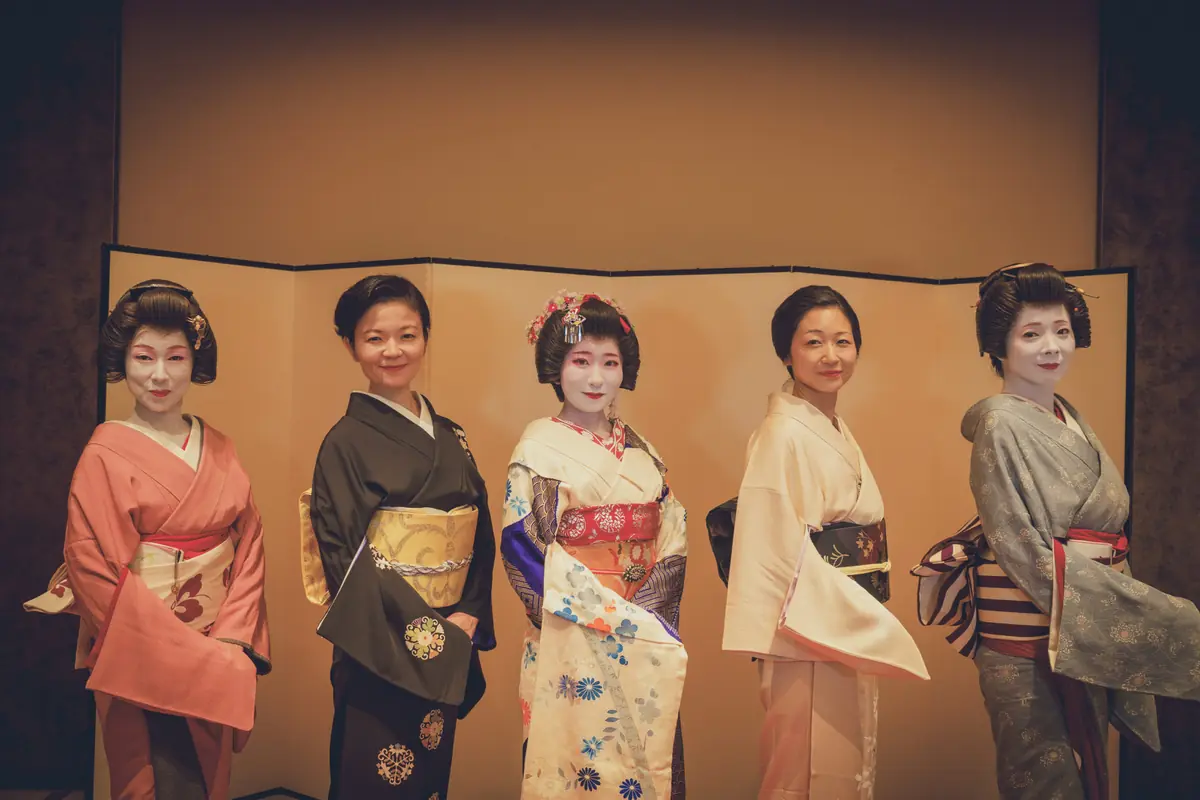 Five women in colorful kimono outfits posing together in front of a gold folding screen in a studio backdrop