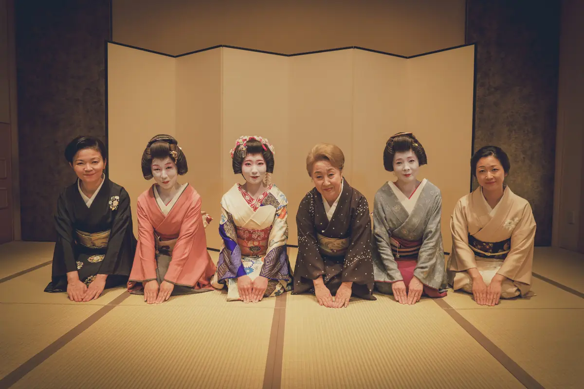 Six women in colorful kimonos kneel on tatami in front of a folding screen, posing for a group photo.