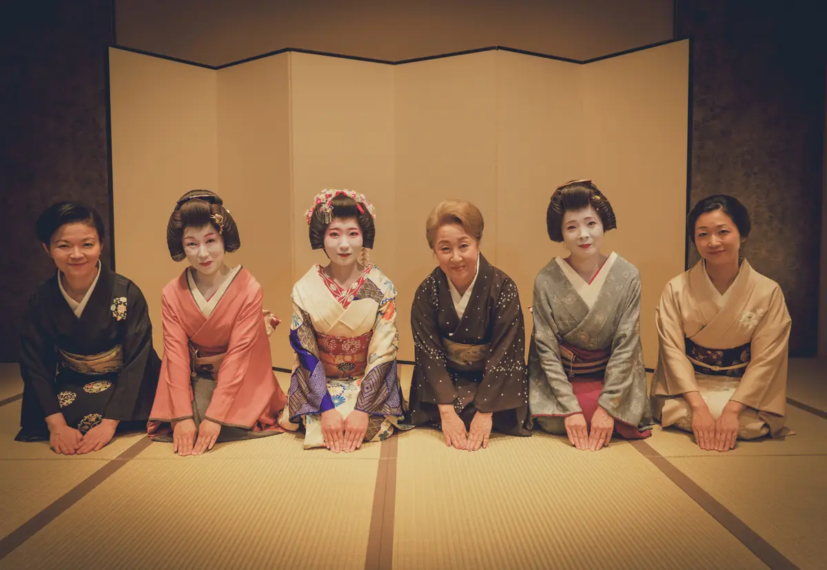 Six women in elaborate kimonos kneeling on tatami mats for a group photo in a traditional Japanese room behind a folding screen.