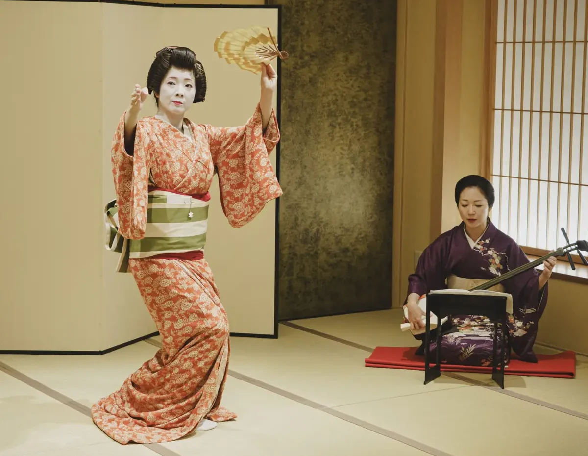Geisha in a pink kimono dances with a fan as a musician in purple plays a shamisen in a tatami room with a folding screen.