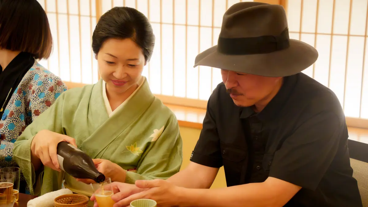 A geisha kneels at the tatami before handing a drink to a guest — eye-level matters in ozashiki etiquette