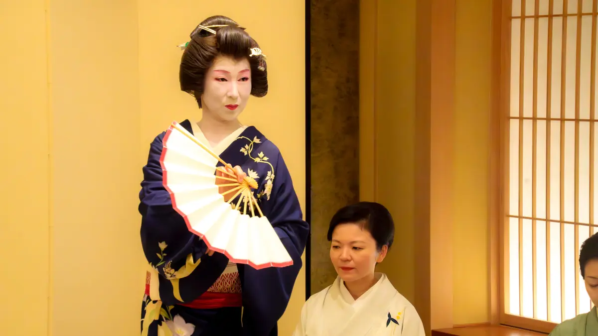 Geisha in a dark blue kimono with floral embroidery stands with a white and red-edged folding fan, beside a seated woman in a white kimono.