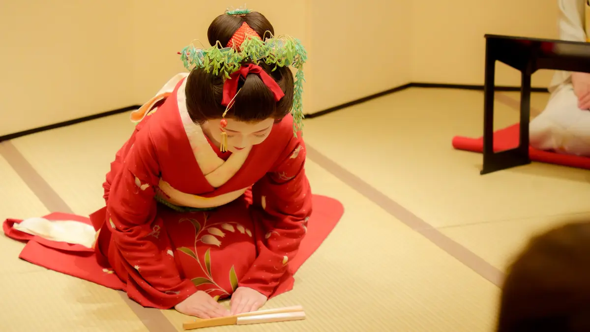 Person in a red kimono with a green leafy headpiece, kneeling on tatami and placing wooden sticks on the floor.