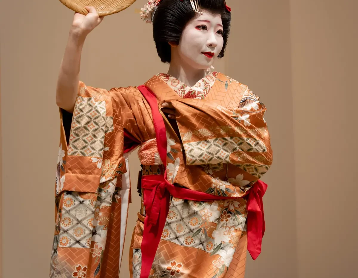 Geisha in an orange patterned kimono with white makeup, raising a round woven hat.