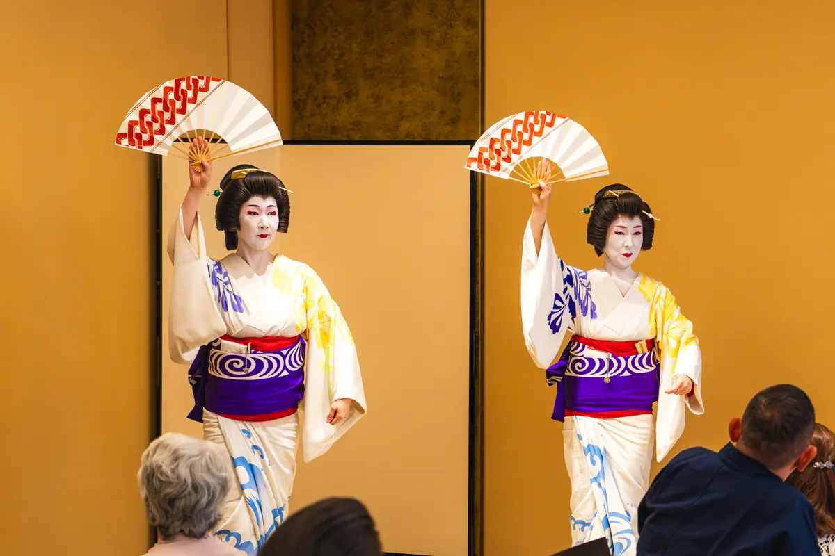 Two geisha dancers in colorful kimonos perform on a gold-stage backdrop, each holding a white fan above