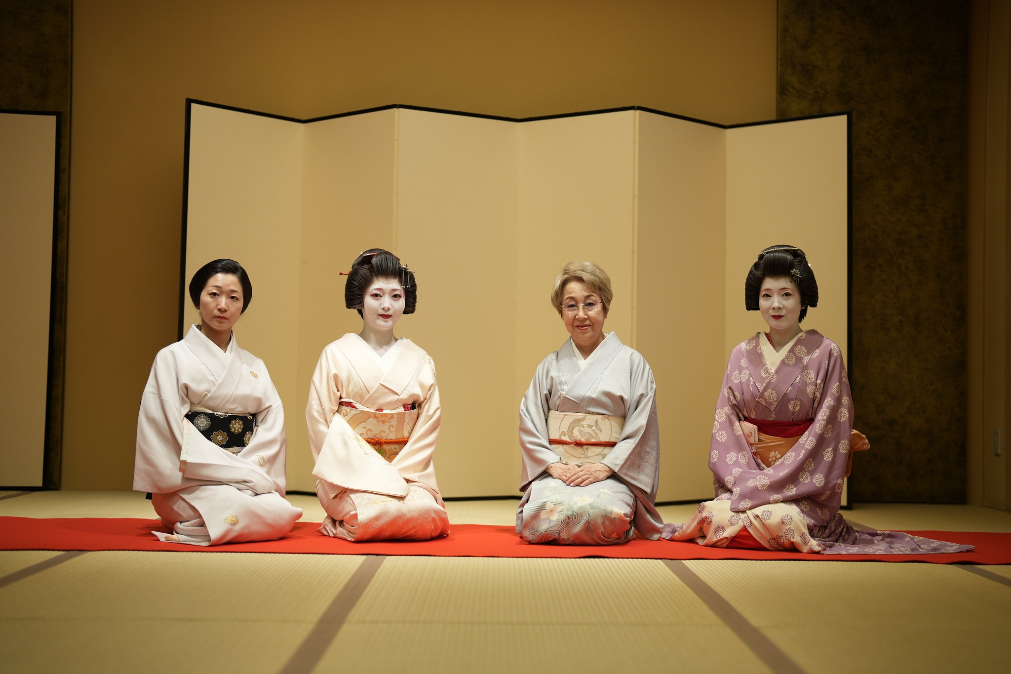 Three Asakusa geisha and the okami gathered for a group portrait in a tatami room with a gold screen