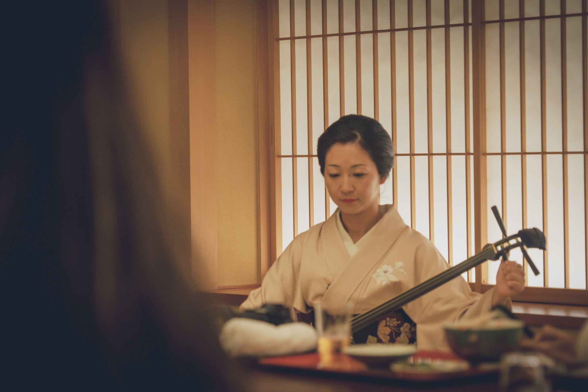 An Asakusa geisha in a cream kimono playing the shamisen beside a latticed window at Miyakodori