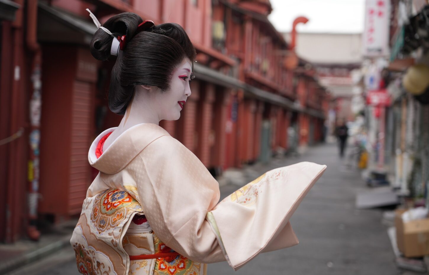 Geisha wearing a pale pink kimono with ornate embroidery, in a side profile on a red-painted street backdrop.