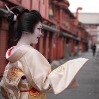 Geisha wearing a pale pink kimono with ornate embroidery, in a side profile on a red-painted street backdrop.