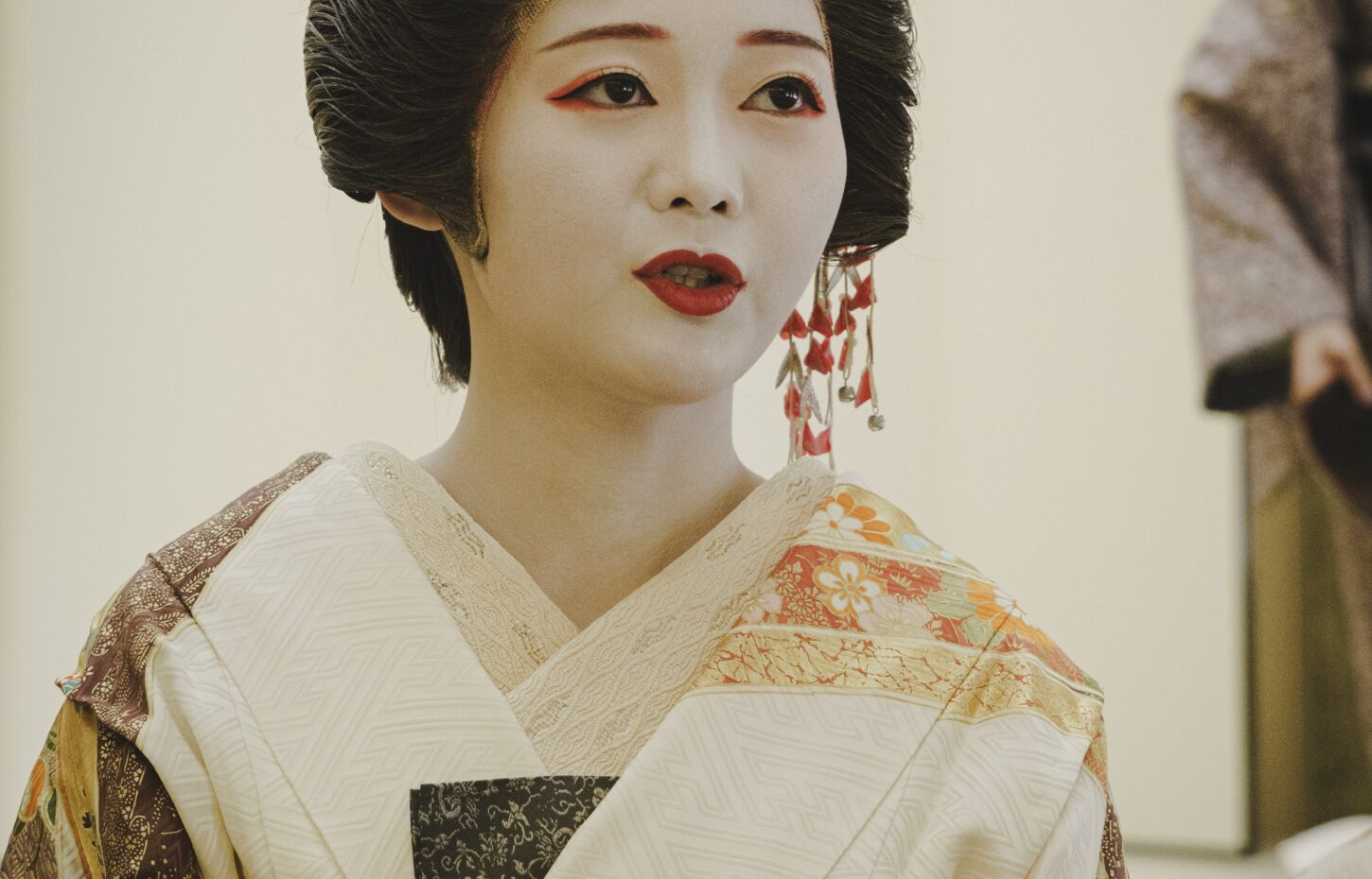 A geisha in a cream and gold silk kimono with floral kanzashi and white makeup