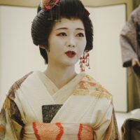 A geisha in a cream and gold silk kimono with floral kanzashi and white makeup