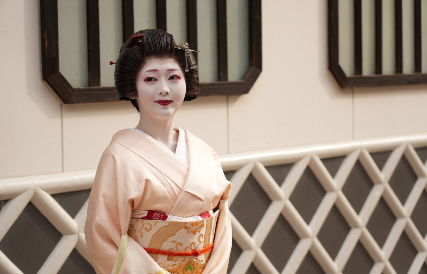 Geisha in a pale pink kimono with an ornate obi, standing against a beige wall with a decorative lattice behind her.