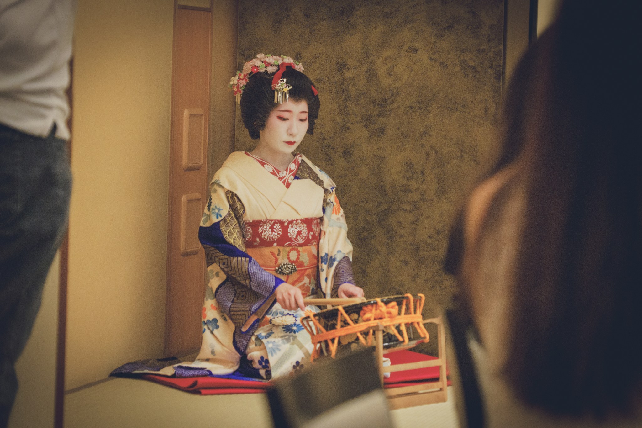 A geisha in floral kanzashi performing with a traditional narimono percussion instrument