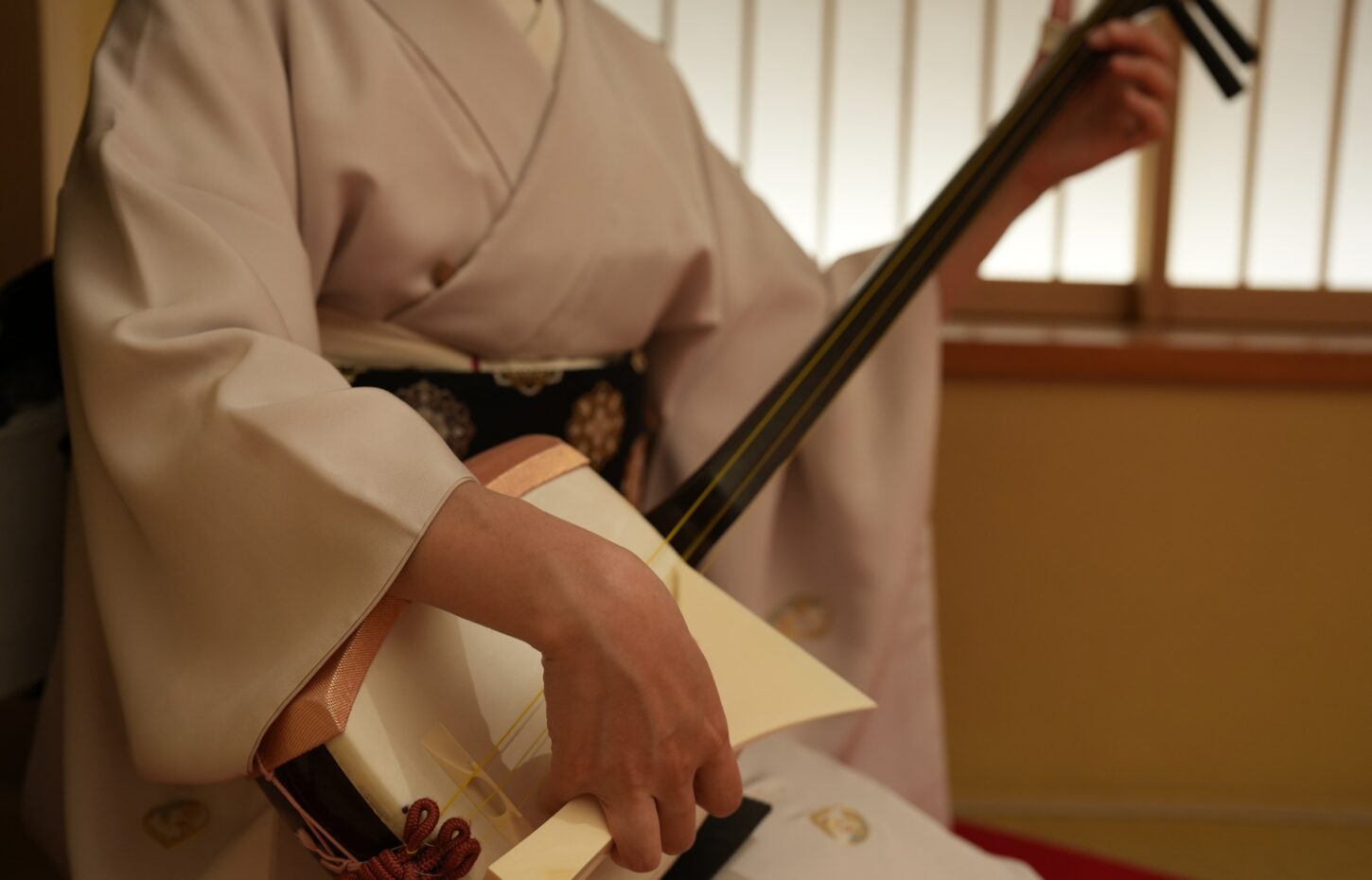 An Asakusa geisha in a white kimono playing the shamisen against a shoji screen — the three-stringed lute at the heart of geisha music