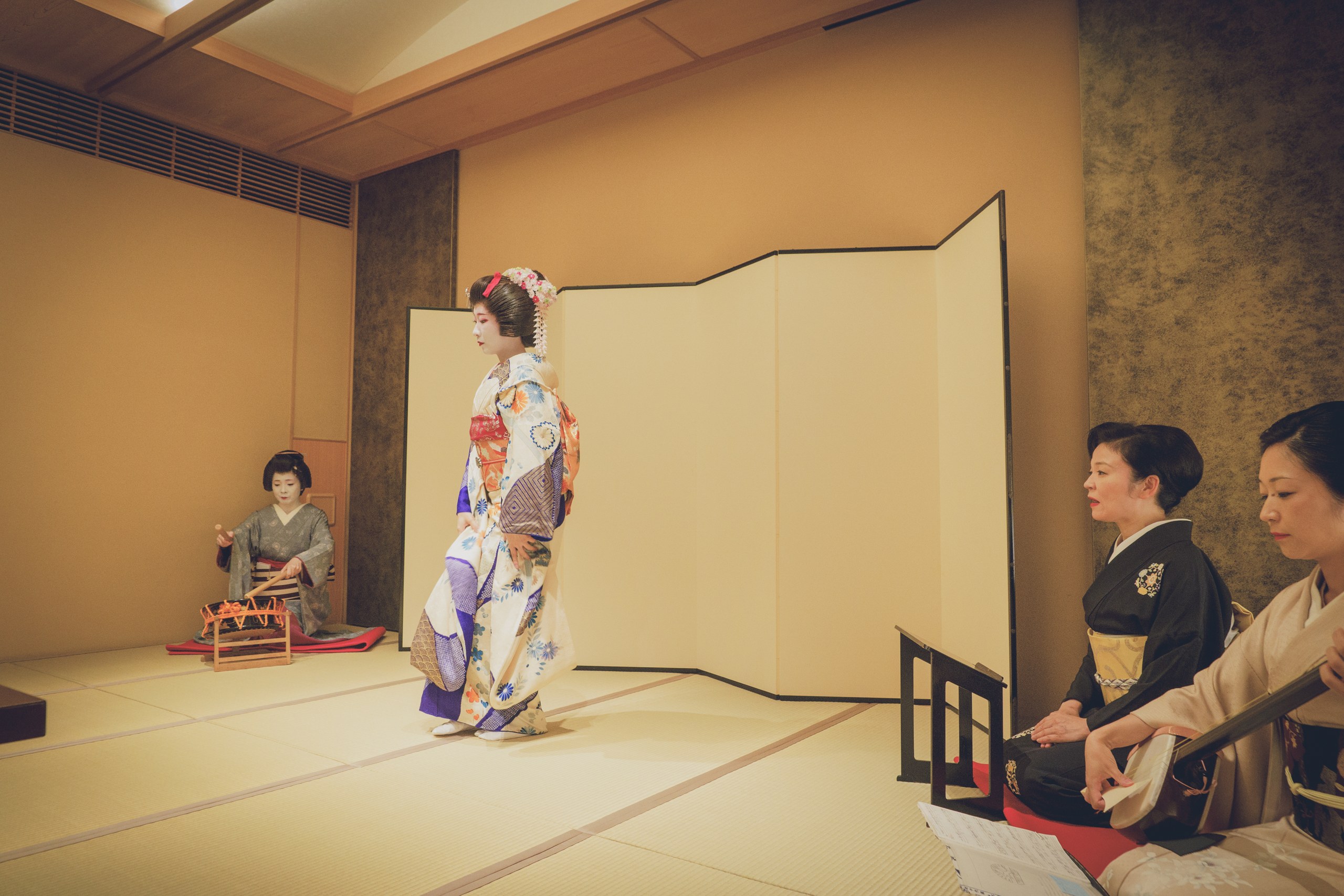 Geisha musicians (jikata) playing narimono percussion while a dancer performs in an ozashiki, against a gold folding screen