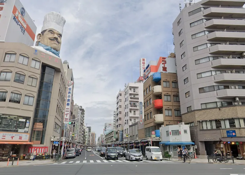 Wide city street in Japan with a giant chef statue on a building to the left; cars, pedestrians, and crosswalks along a lined urban avenue.