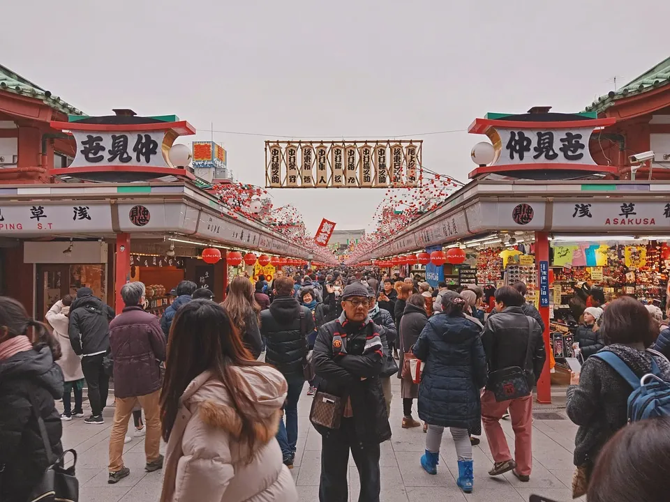 Crowded street market with stalls, red lanterns, and Japanese signage shoppers browsing under a decorative archway.