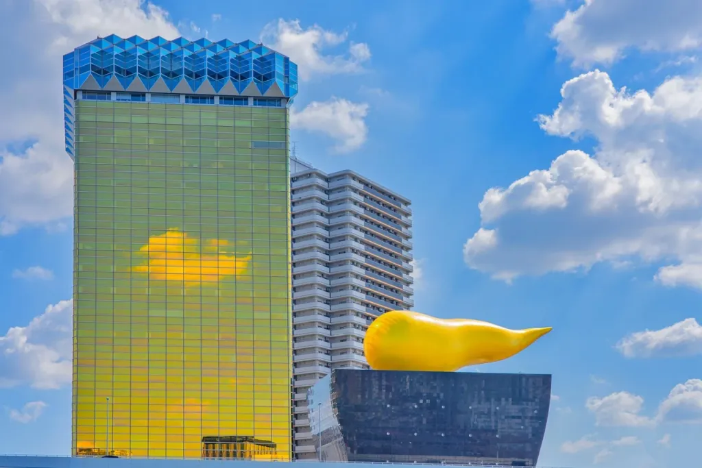 Urban skyline with a tall glass skyscraper reflecting a golden sunset and a large yellow sculpture atop a neighboring dark podium building,