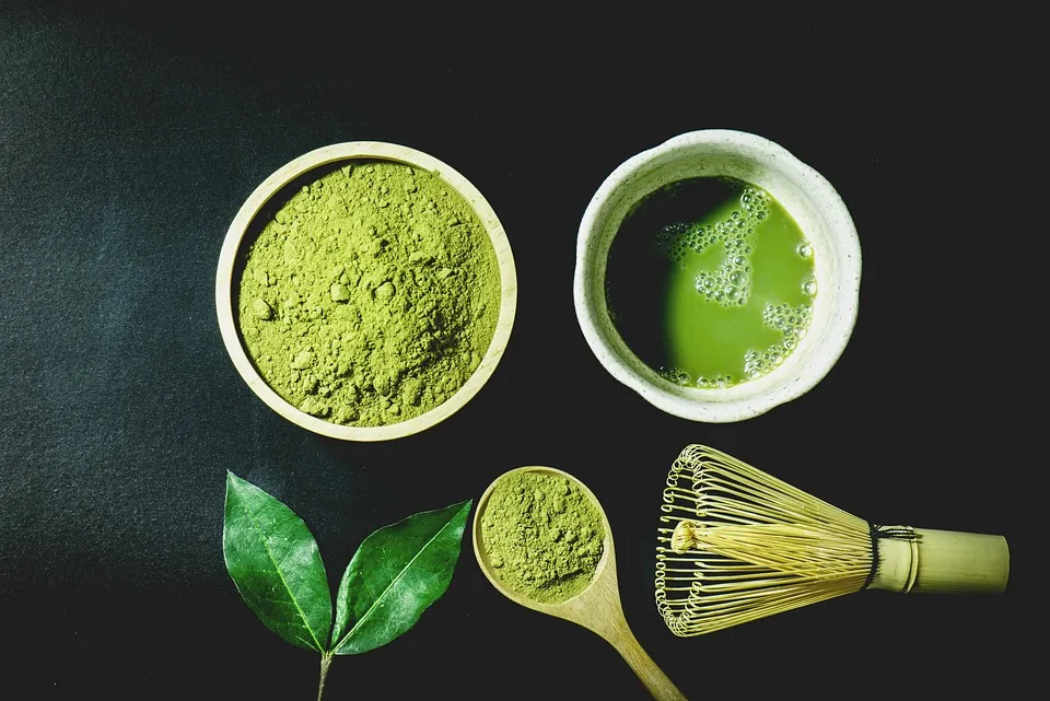 Top-down view of matcha tea ceremony tools: bowls of green powder, a bamboo whisk, a spoon, and fresh leaves on a dark surface.