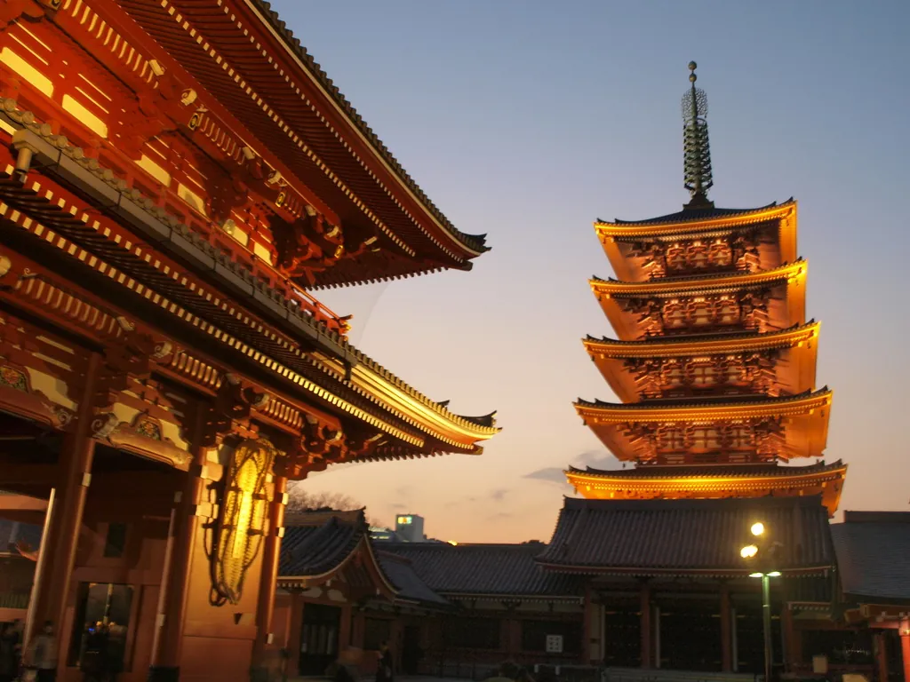 Five-story pagoda lit with warm lights beside a traditional temple at dusk.