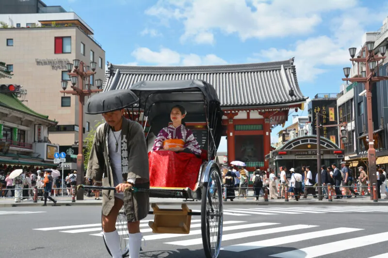 A rickshaw being pulled by a man in traditional clothing carries a woman in a red kimono past a temple gate on a busy street lined with shops and pedestrians.