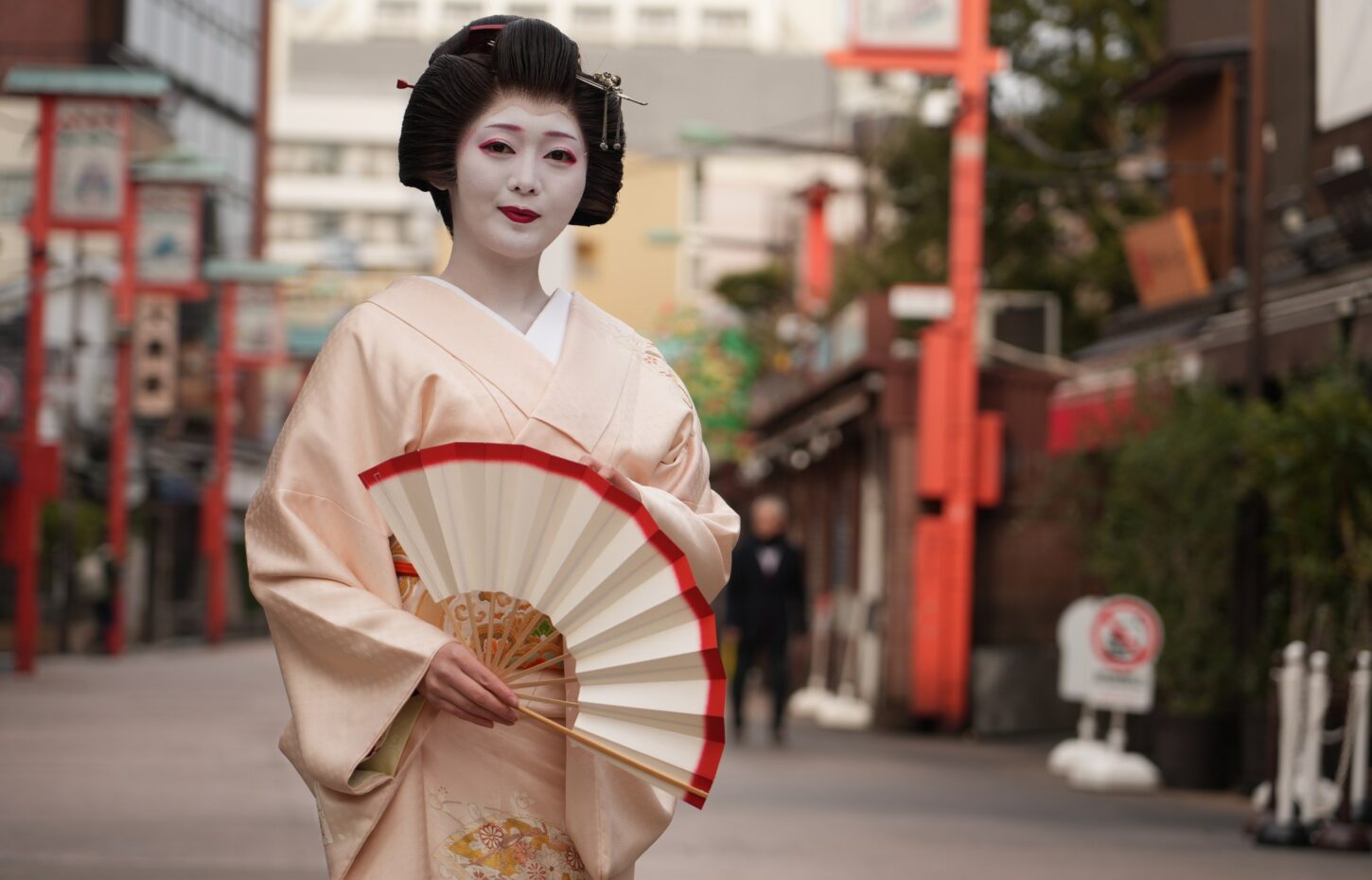 Geisha in a pale peach kimono with white makeup and red lips, holding a white and red folding fan outdoors.