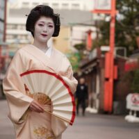 Geisha in a pale peach kimono with white makeup and red lips, holding a white and red folding fan outdoors.