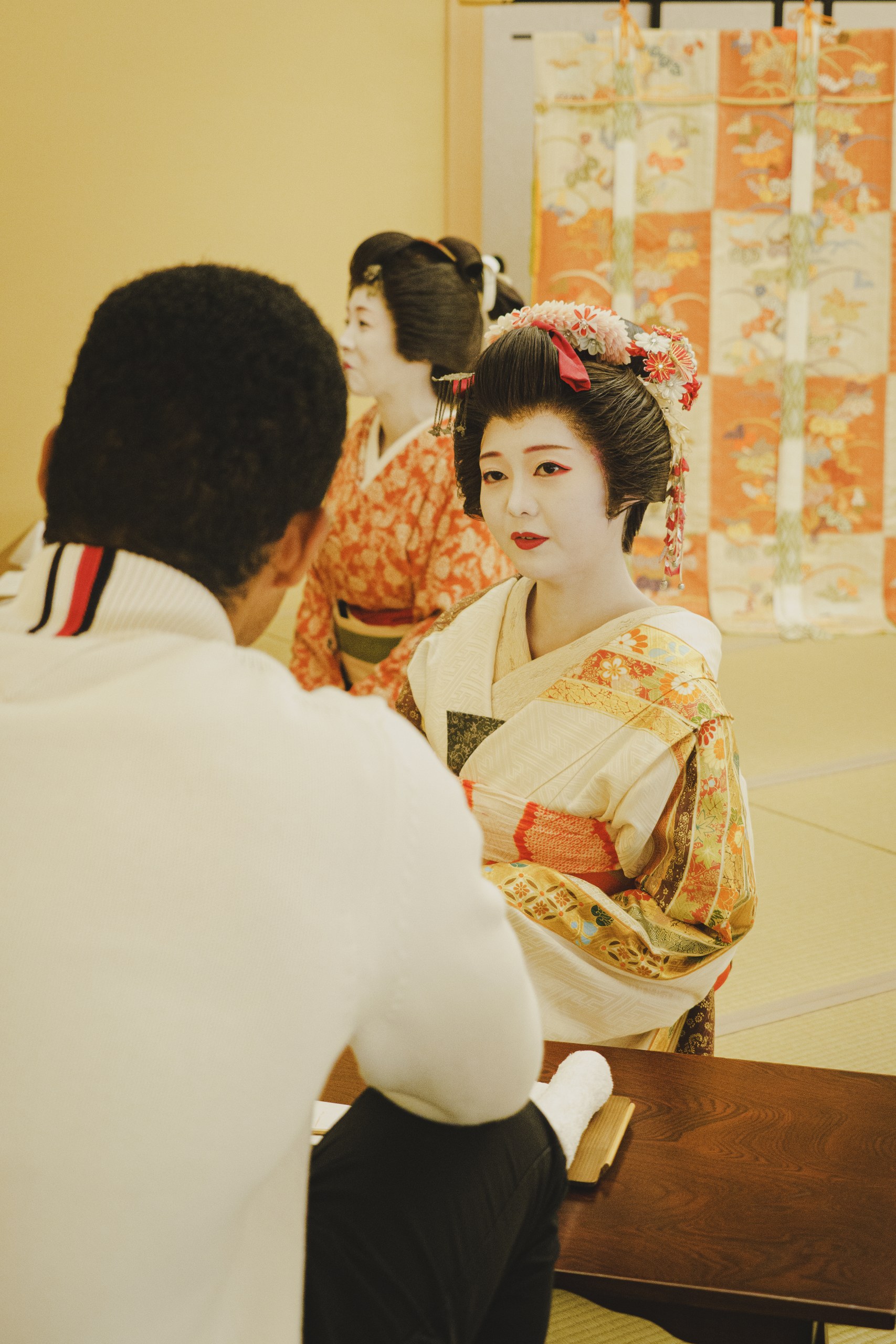 Two Asakusa geisha in full kimono with a foreign guest at the Ozashiki Tea House at Miyakodori, against a gold folding screen — a commemorative moment