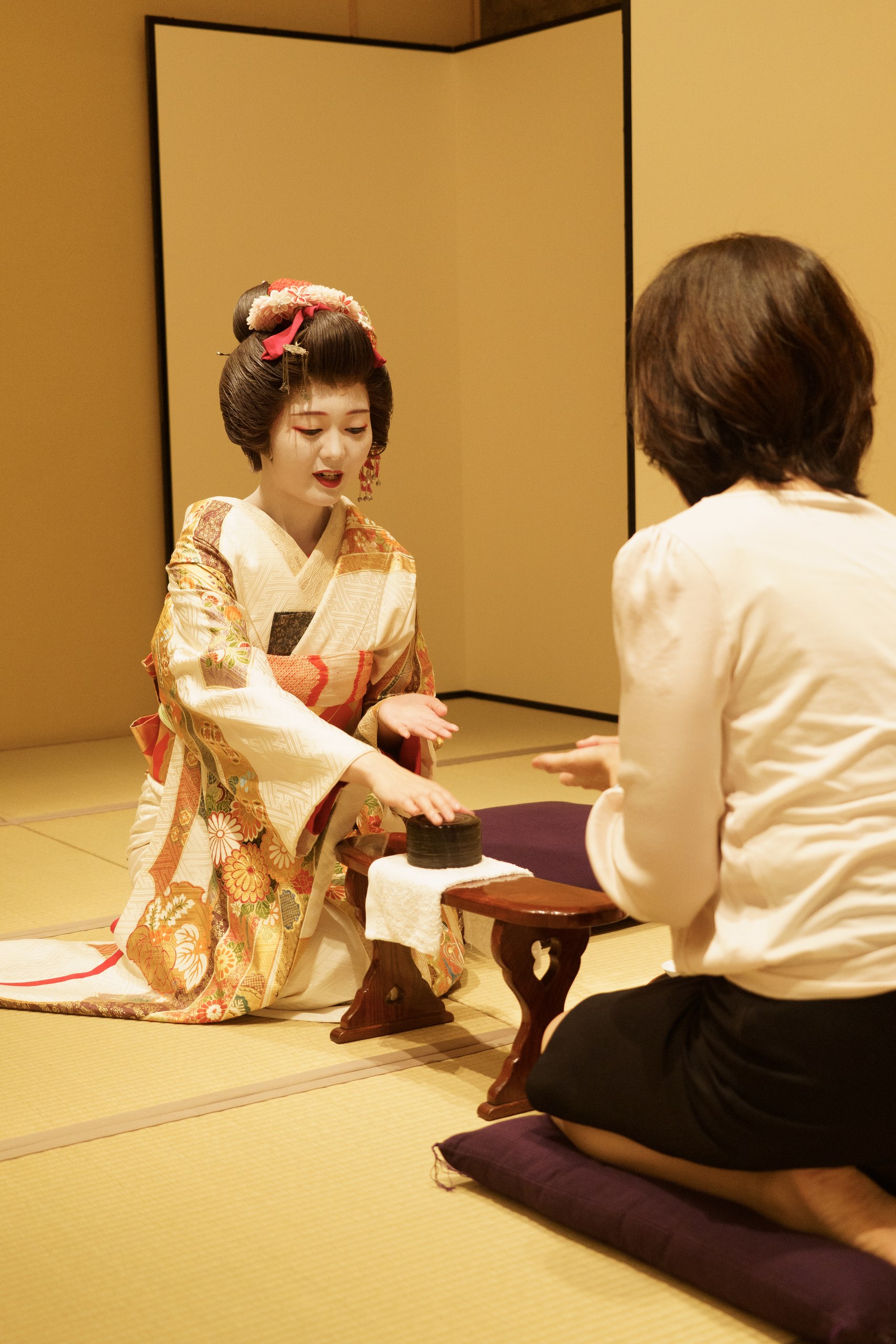 An Asakusa geisha presenting seasonal wagashi to a guest at the Ozashiki Tea House at Miyakodori — matcha and sweets served as part of an afternoon with working geisha