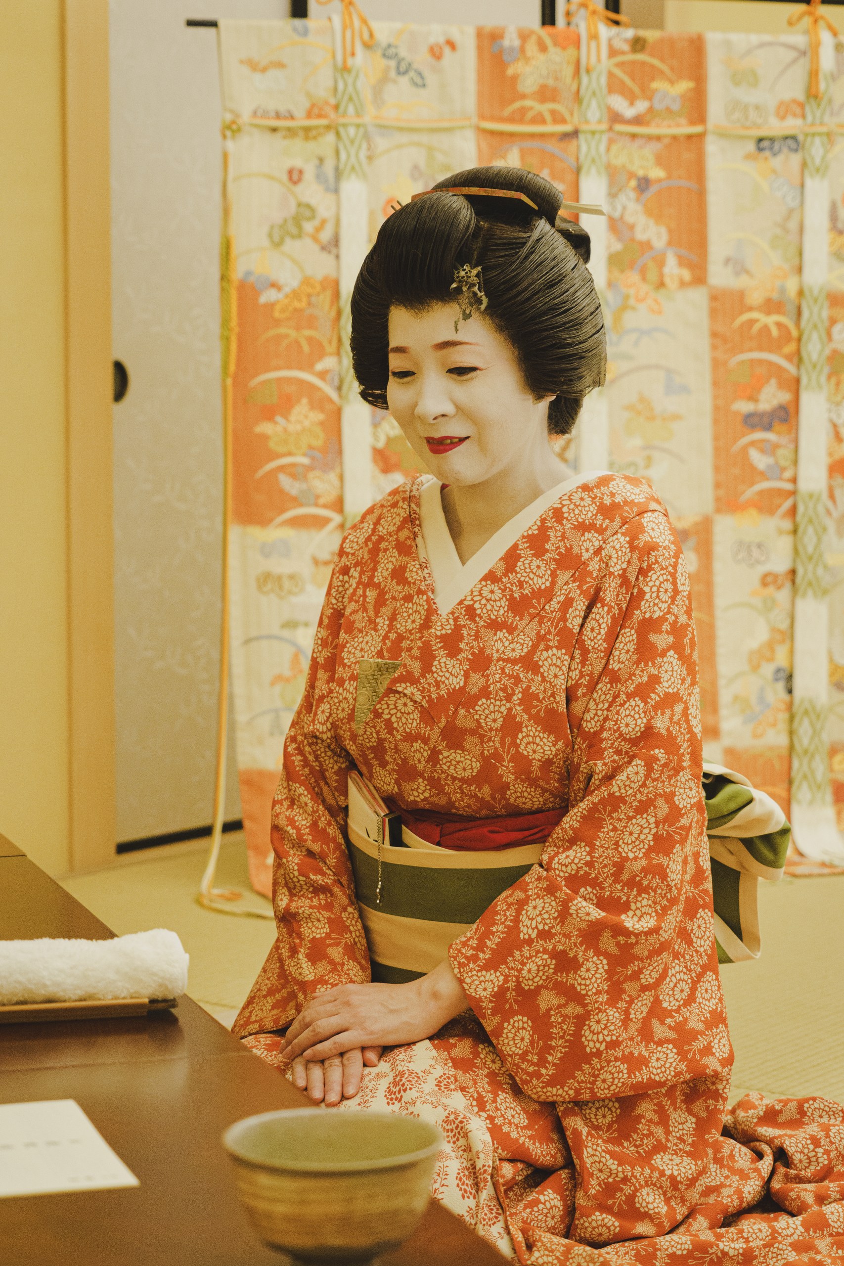 An Asakusa geisha in a red komon kimono with a bowl of matcha green tea, in a warmly lit tatami room at Miyakodori