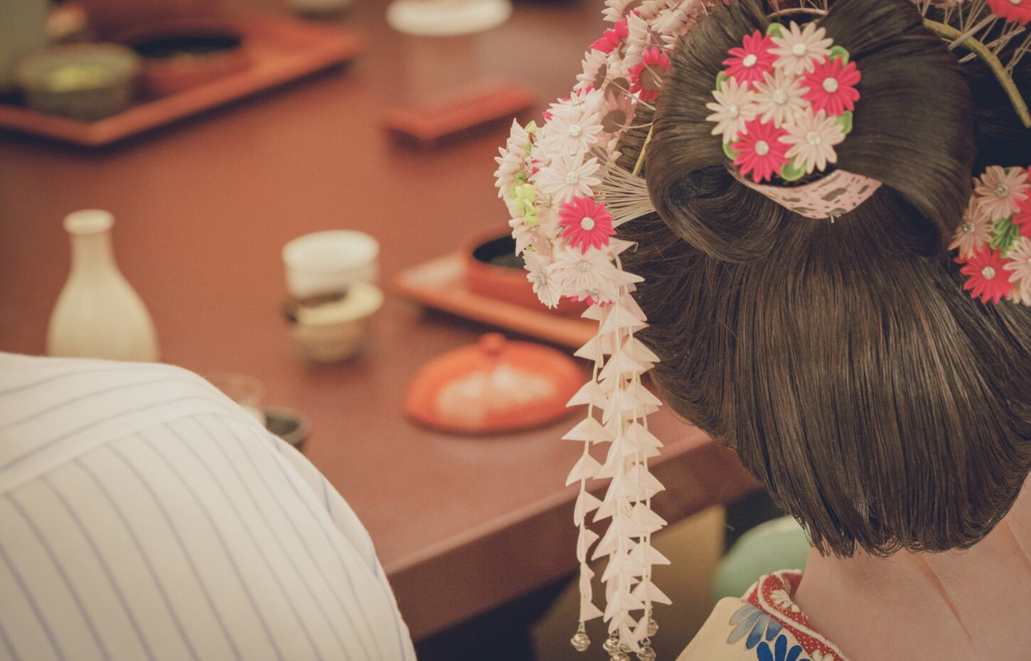 Back of a person’s head with dark hair wearing pink and white flower hair accessories at a festival/ceremony orientation.