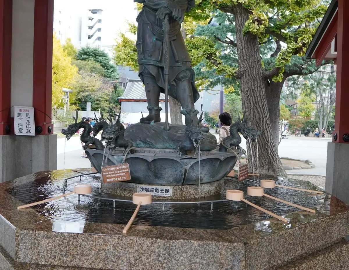 Bronze armored warrior statue atop a dragon-themed fountain in a red pavilion, water spouting around it.