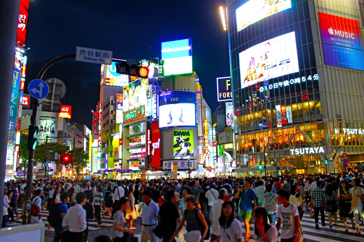 Crowded crosswalk at night in Shibuya, Tokyo, with neon billboards and a glass-front building towering over pedestrians earlier in the evening.