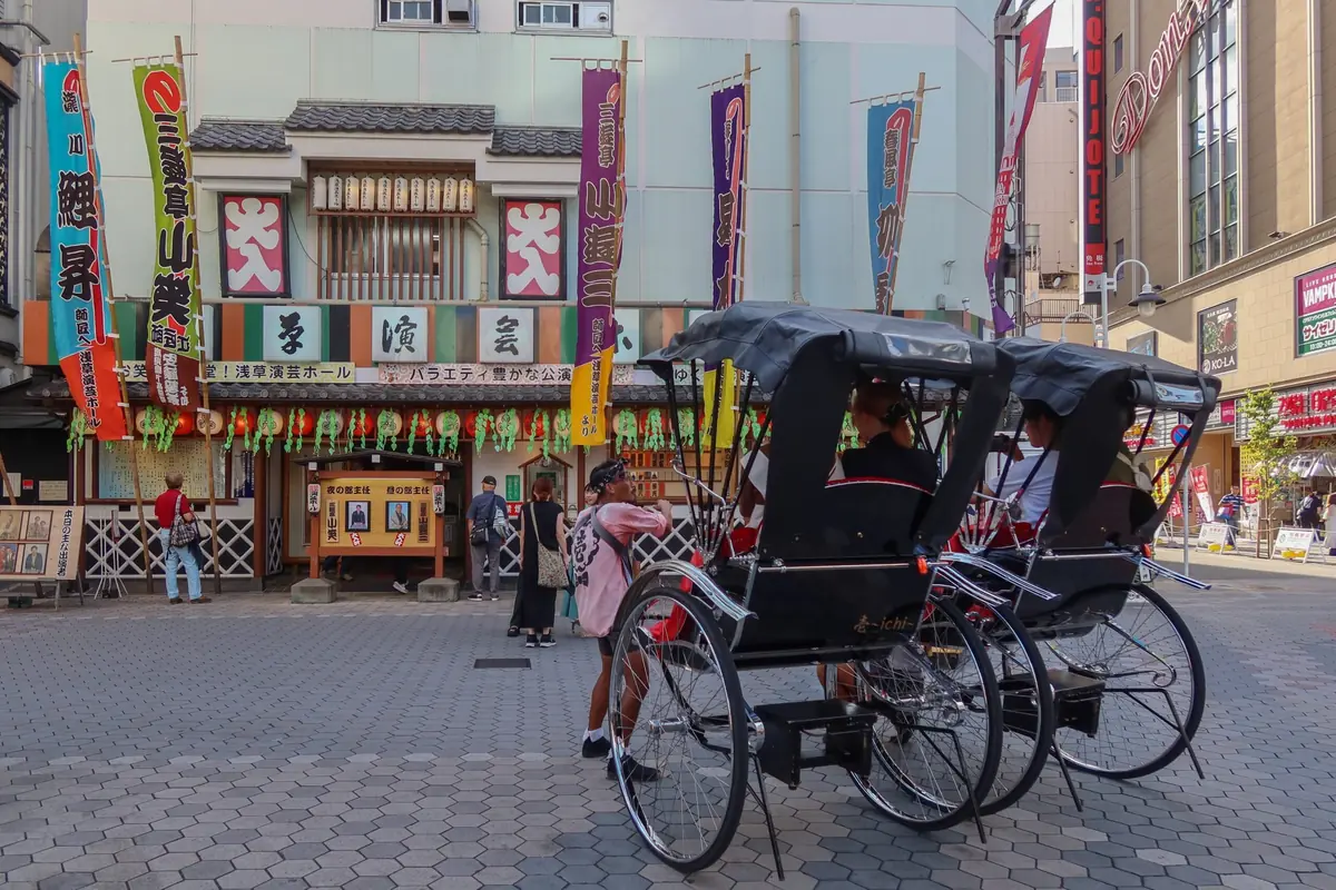 Two-wheeled rickshaw parked on a patterned plaza in front of a colorful Japanese theater facade with vertical banners.