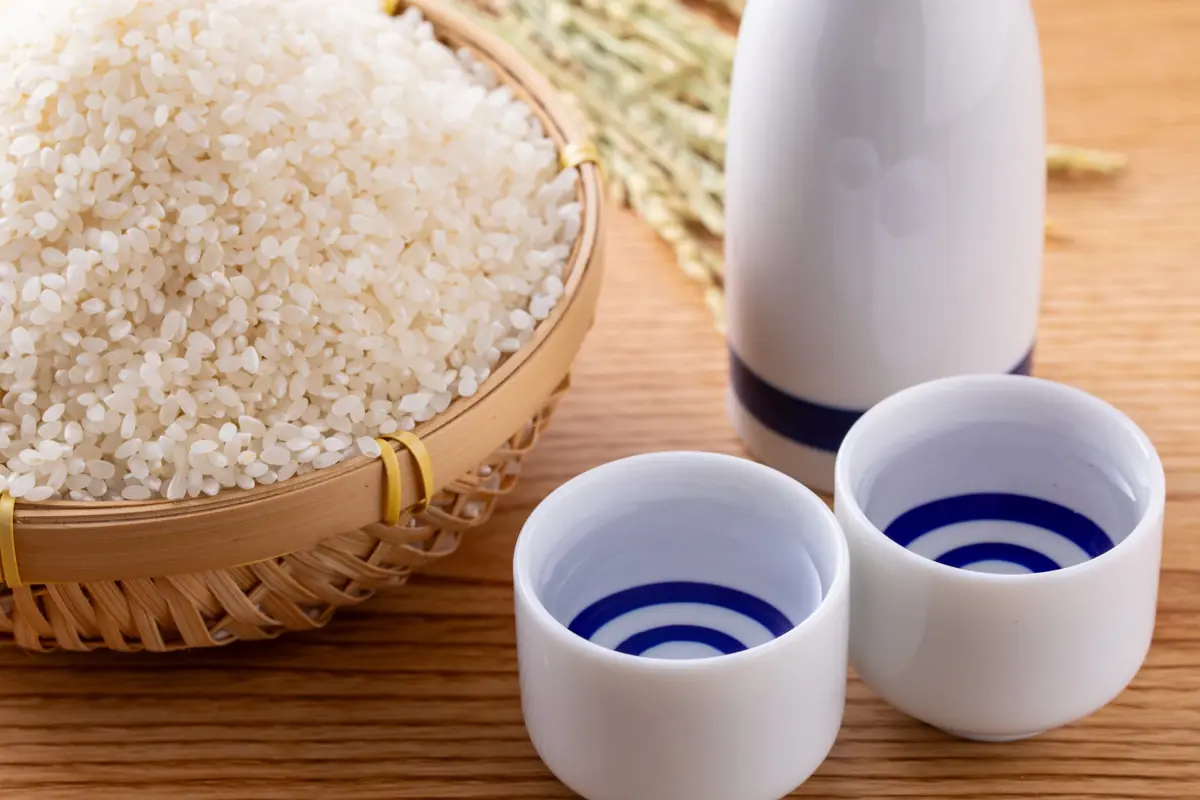 Basket of white rice beside a white sake bottle and two small cups on a wooden surface, suggesting a Japanese meal setup.