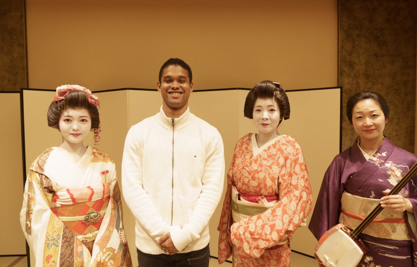 Four people pose in traditional Japanese attire: a smiling man in a white sweater flanked by three women in colorful kimonos, one holding a shamisen instrument