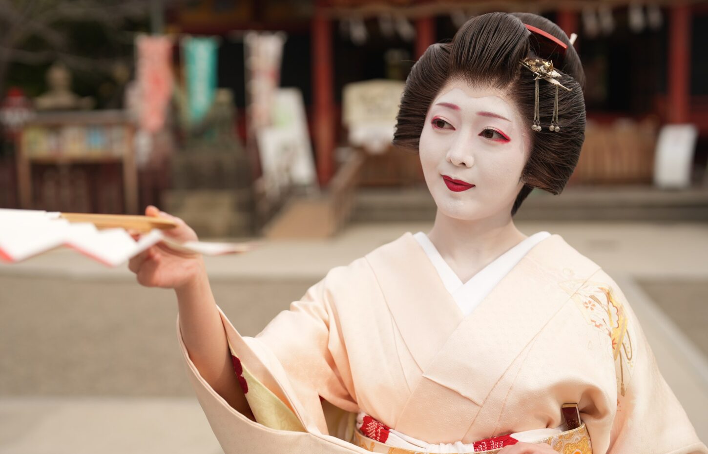 Geisha with white makeup and red lips in a pale kimono, extending papers toward the camera.