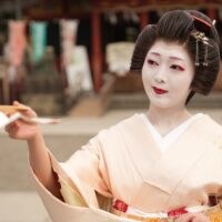 Geisha with white makeup and red lips in a pale kimono, extending papers toward the camera.