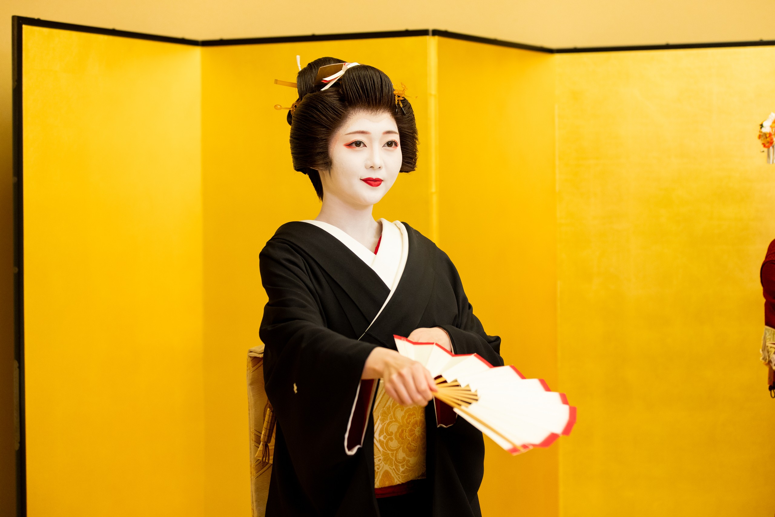 Geisha in black kimono presenting an open fan before a golden folding screen
