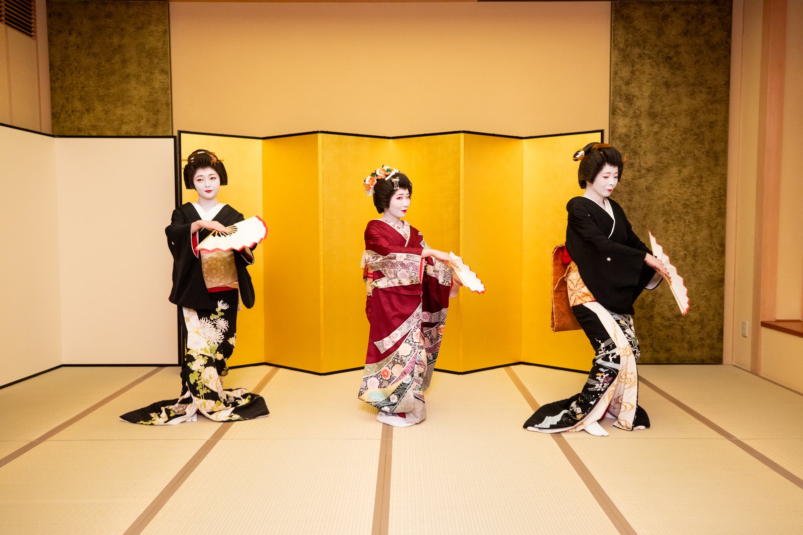 Three Asakusa geisha dancing together with folding fans in front of a gold screen