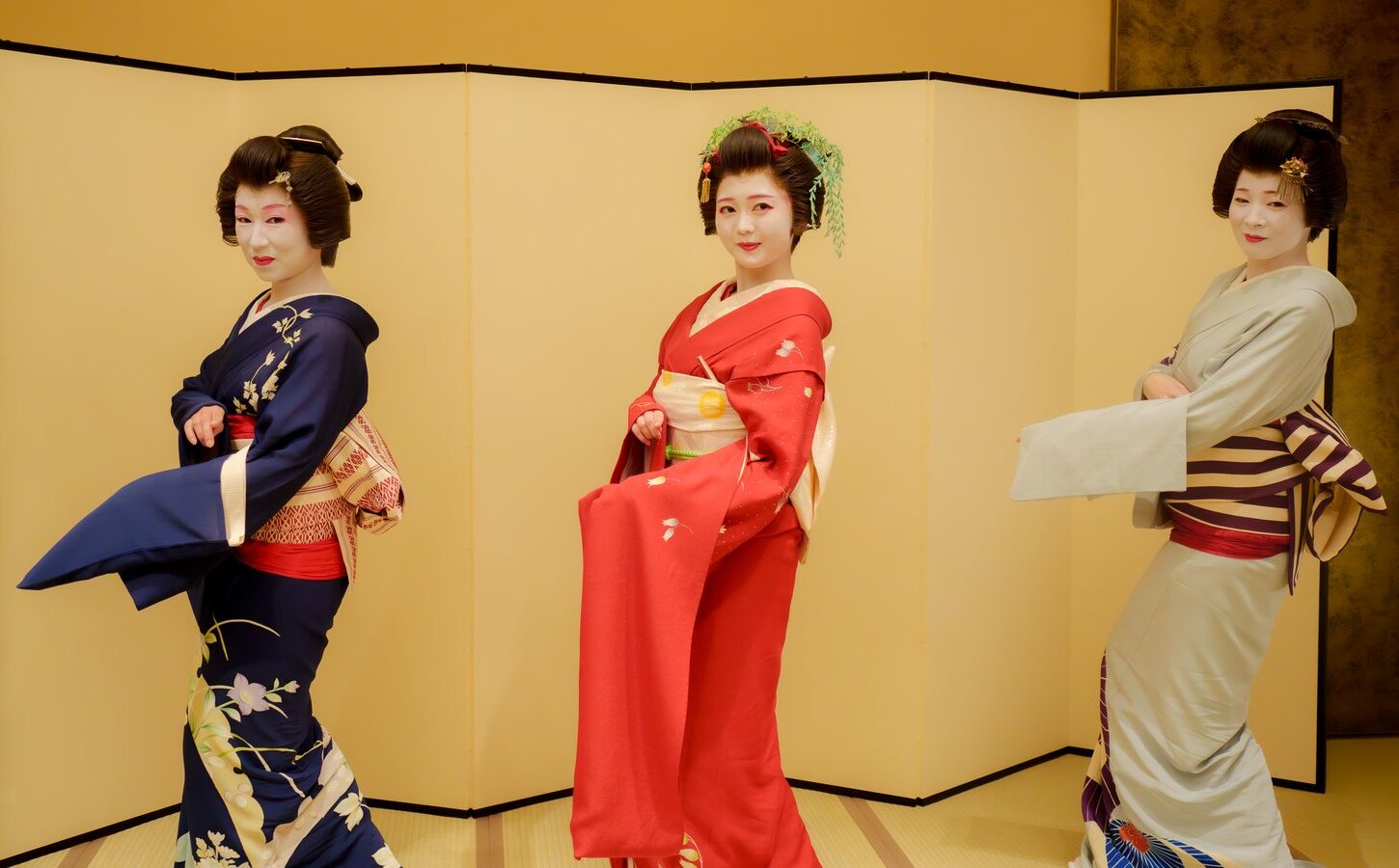 Three Asakusa geisha posed in front of a folding screen, a hangyoku in red furisode between two ippon geisha
