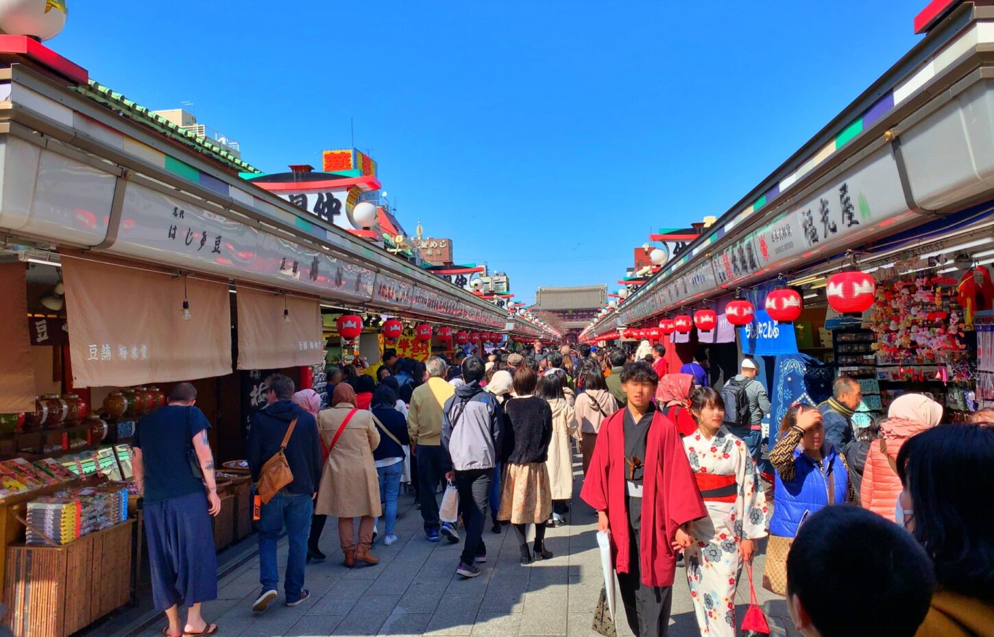 A couple in kimono walk along Nakamise-dori in Asakusa, lined with shops and red lanterns, with Senso-ji's Hozomon gate ahead