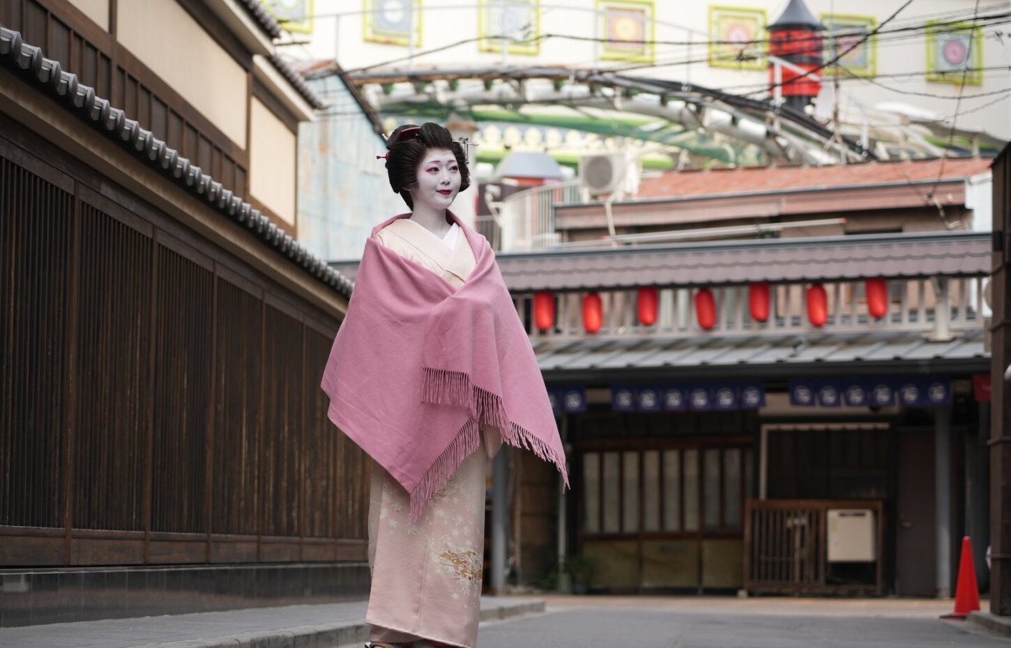 Geisha in a pale pink kimono with a matching shawl, standing on a quiet street lined with wooden walls and red lanterns in the background.