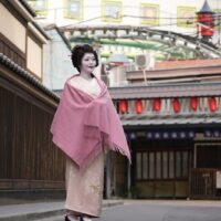 Geisha in a pale pink kimono with a matching shawl, standing on a quiet street lined with wooden walls and red lanterns in the background.