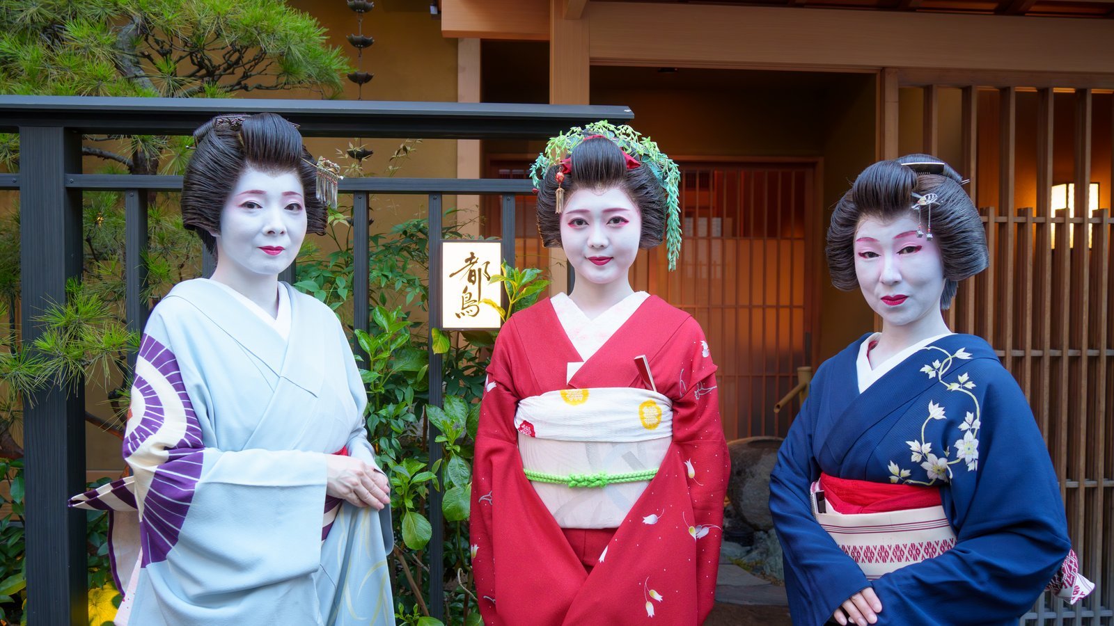 Three Asakusa geisha standing outside the entrance of Miyakodori, a machiai-chaya in Asakusa, beside the illuminated house sign