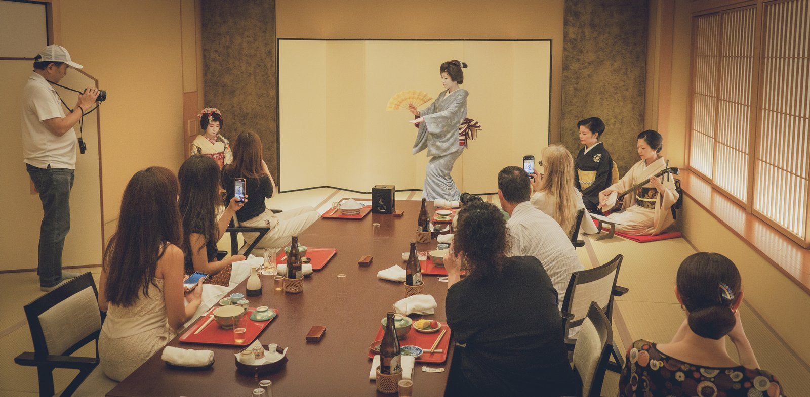 A geisha dancing to live shamisen music in front of seated guests at a Miyakodori ozashiki