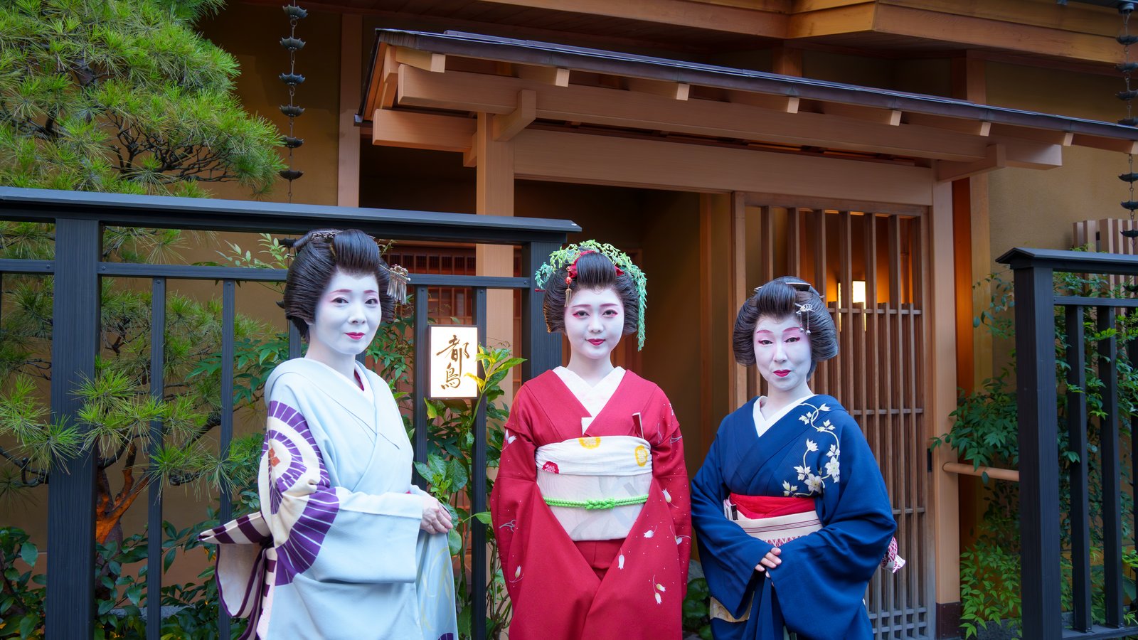 Three Asakusa geisha standing at dusk outside Miyakodori, beside the lantern sign of the machiai-chaya that takes bookings without an introduction