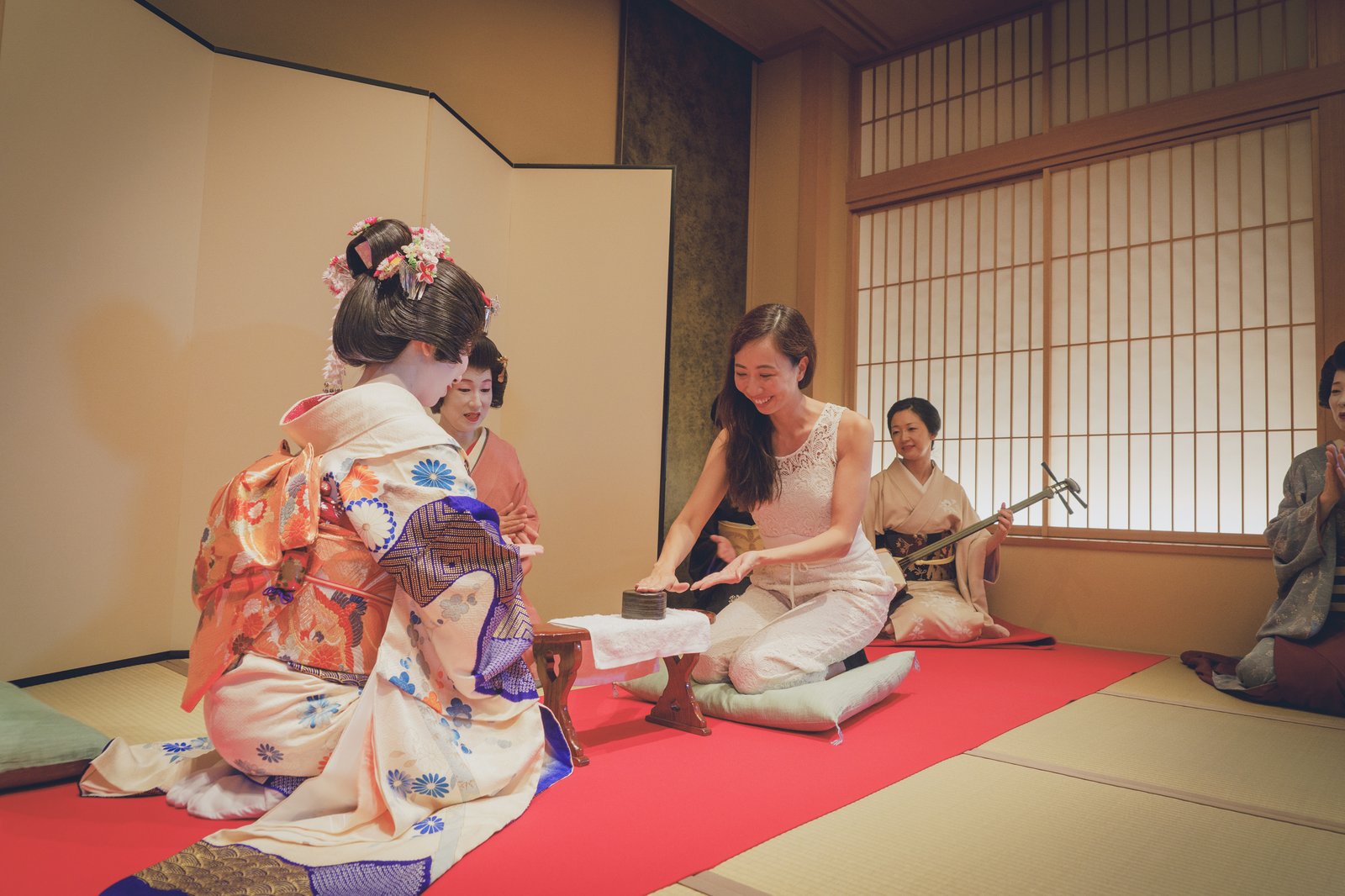A guest playing Konpira Fune Fune with a hangyoku across a small wooden stand at a Miyakodori ozashiki, with a jikata on shamisen behind them