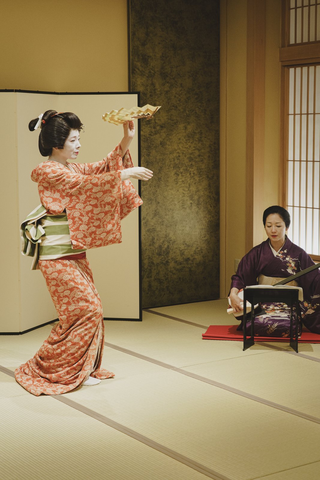 An Asakusa geisha mid-dance, her obi tied at the back, beside a seated shamisen player