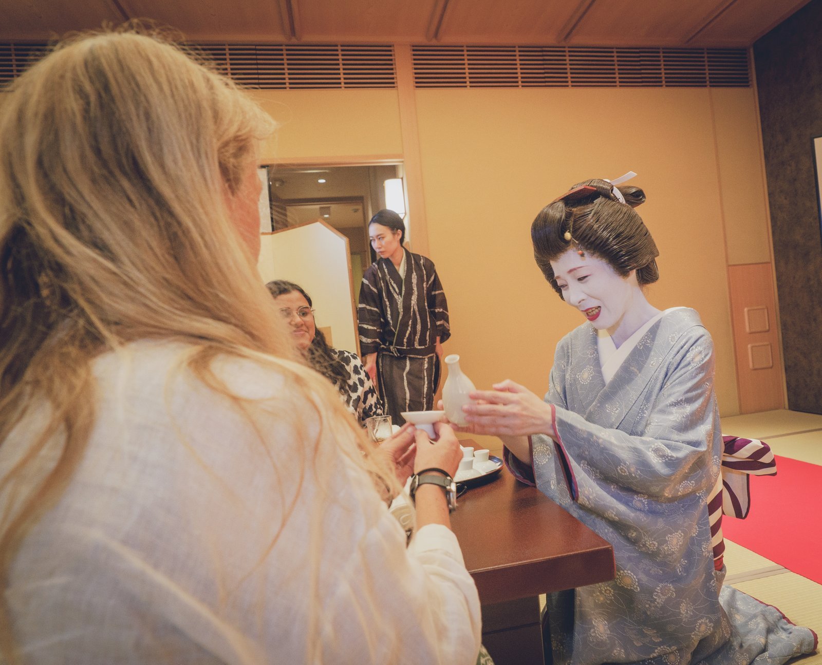 A geisha in silk kimono kneeling to pour sake for a guest at a low table in a private tatami room